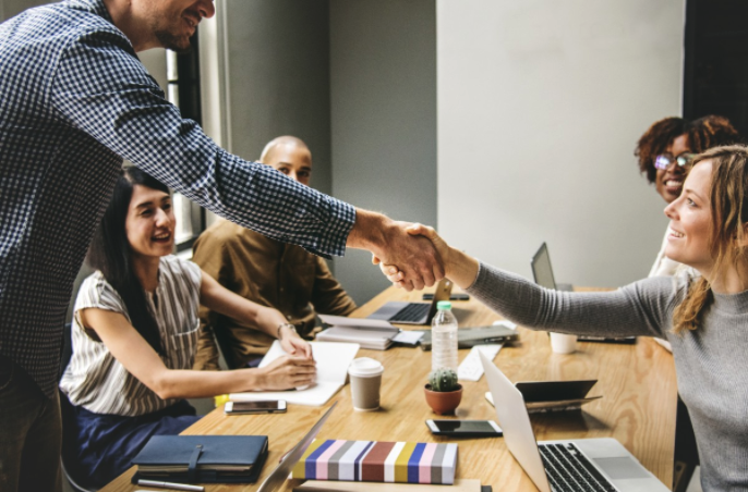 A male and a female shaking hands as a b2b sales agreement, over the table with their teams sitting around