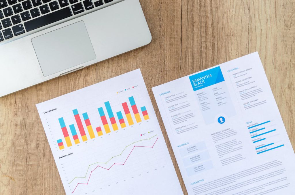 a resume and a chart on a desk next to a laptop, as a representation of uploading resume and certificates to linkedin