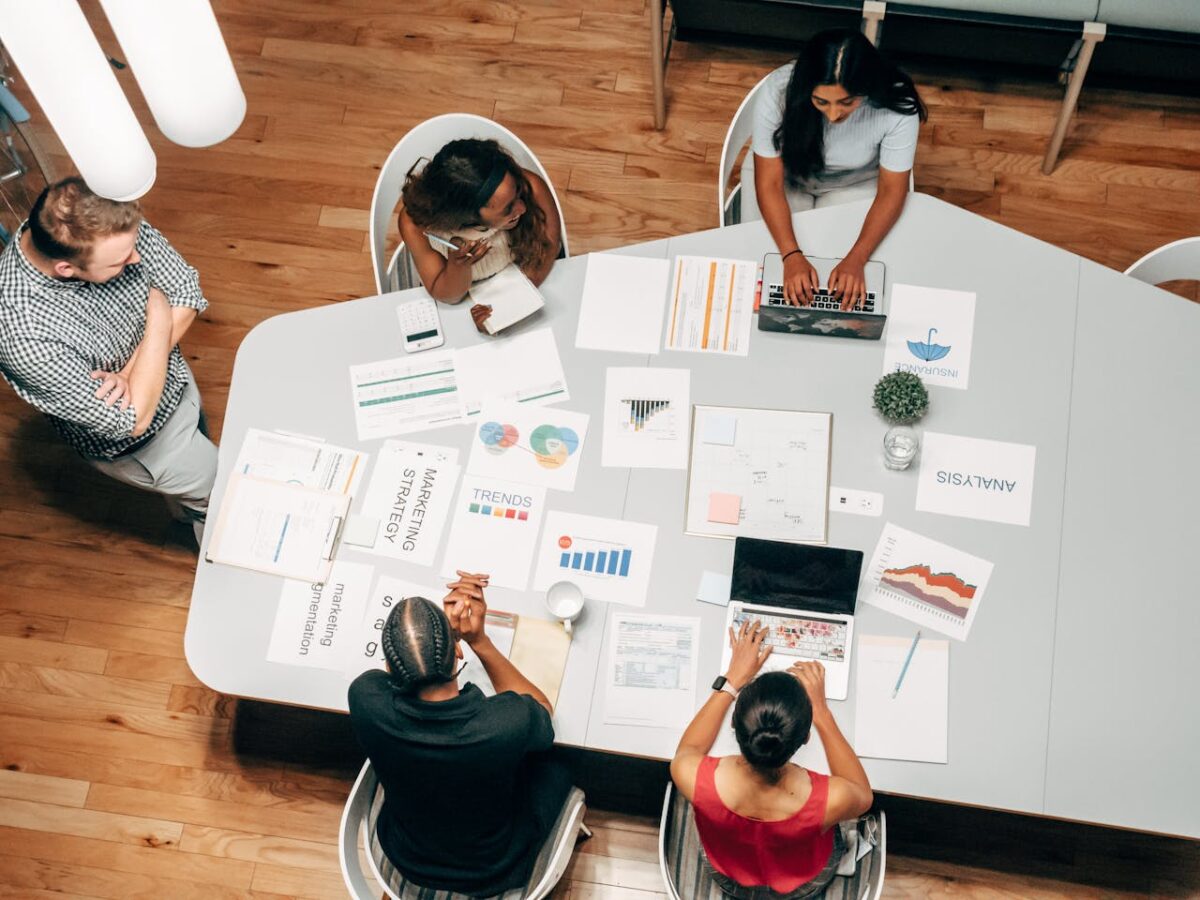 Five employees at the meeting table looking at charts and graphs while working on laptops.
